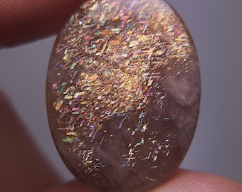 May include: Oval-shaped sunstone gemstone with a shimmering, iridescent effect. The stone displays a mix of colors, including gold, pink, and purple, with a speckled pattern. The gemstone is held between two fingers.