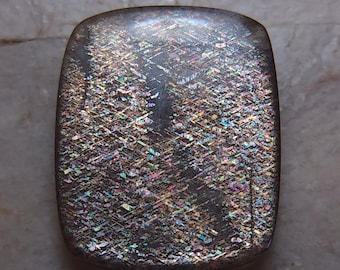 May include: A dark gray, rectangular gemstone with iridescent rainbow colors. The gemstone has a textured surface with a pattern of lines.