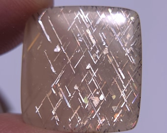 May include: A square-shaped, translucent, light pink gemstone with a silver and gold metallic pattern. The gemstone has a shimmering, iridescent effect.