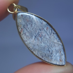 May include: A gemstone pendant with a gold-colored setting. The pendant has a clear, elongated shape with a pattern of gray, black, and reddish-brown inclusions. The pendant is held by a finger.