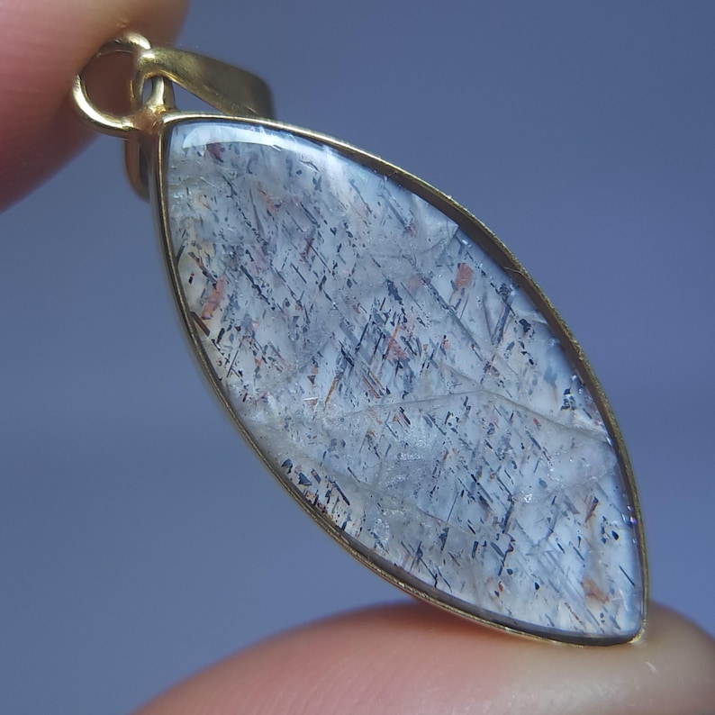 May include: A gemstone pendant with a gold-colored setting. The pendant has a clear, elongated shape with a pattern of gray, black, and reddish-brown inclusions. The pendant is held by a finger.