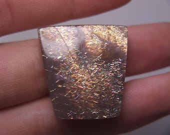 May include: A rectangular sunstone gemstone with a polished surface. The stone displays a shimmering effect with flashes of gold, orange, and iridescent colors. The gemstone is held in a hand.