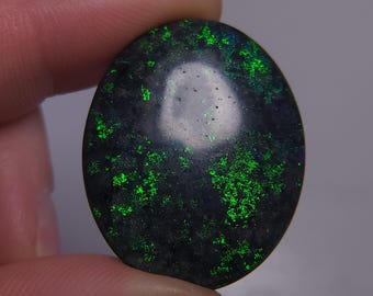 May include: Oval black opal gemstone with bright green flashes. The polished surface reflects light, displaying the play-of-colour effect. The gemstone is held between fingers, highlighting its size and shape.