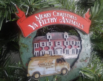 Può includere: Un ornamento natalizio verde e rosso con una casa e un furgone con il testo "Merry Christmas Ya Filthy Animals" e "Oh-Kay Plumbing & Heating"