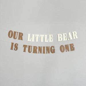 May include: A banner with the text "OUR LITTLE BEAR IS TURNING ONE" in brown and cream-colored letters. The banner is strung on a thin white string and is against a light gray background. Perfect for a first birthday celebration.