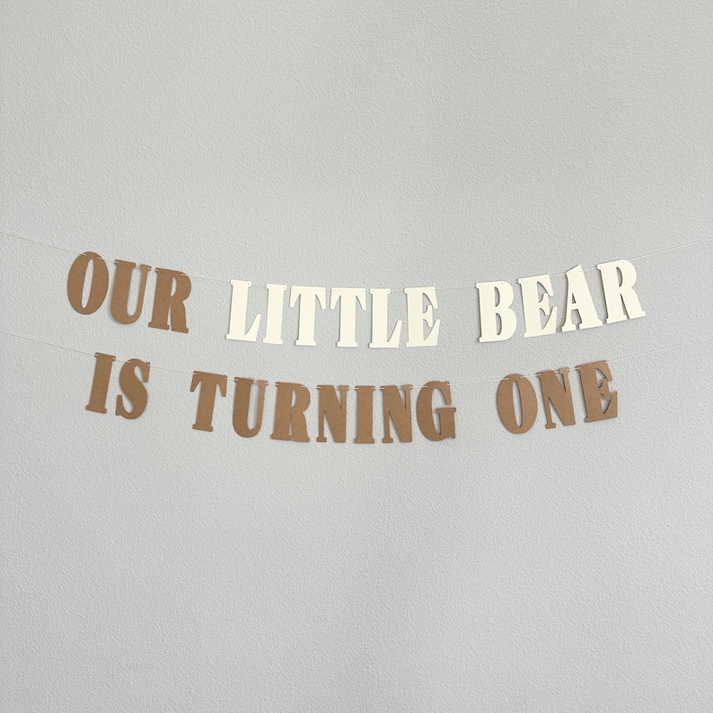 May include: A banner with the text "OUR LITTLE BEAR IS TURNING ONE" in brown and cream-colored letters. The banner is strung on a thin white string and is against a light gray background. Perfect for a first birthday celebration.