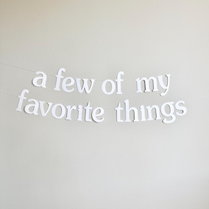 May include: White paper banner with the phrase "a few of my favorite things" strung across a neutral wall. The letters are in a simple, clean font, creating a minimalist aesthetic. The banner is suspended by a thin, nearly invisible string.