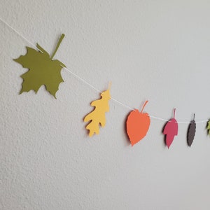 May include: A string of paper leaves in various fall colors, including green, yellow, orange, red, and brown, is strung together with white thread. The leaves are cut out in different shapes, including maple, oak, and heart.