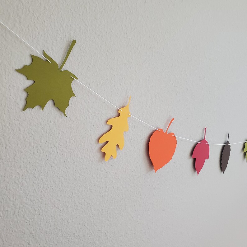 May include: A string of paper leaves in various fall colors, including green, yellow, orange, red, and brown, is strung together with white thread. The leaves are cut out in different shapes, including maple, oak, and heart.