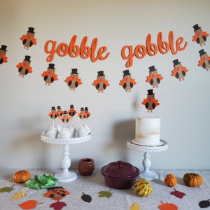 May include: Gobble Gobble banner with orange and brown turkeys, cupcakes with turkey toppers, a white cake with a brown top, and a brown casserole dish on a white table with fall decorations.
