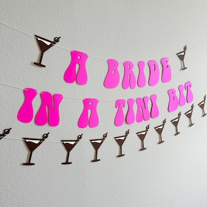 May include: A pink and white paper banner with the text "A Bride in a Tini Bit" and brown paper cutouts of martini glasses.