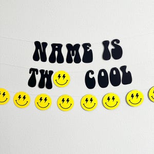 May include: Black and yellow banner with the text "NAME IS TWO COOL" in a retro font. The banner is decorated with yellow smiley faces with lightning bolts for eyes.