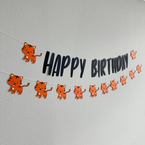 May include: A black and white banner with the text "Happy Birthday" and orange tiger cutouts. The banner is strung with white string and is hanging on a white wall.