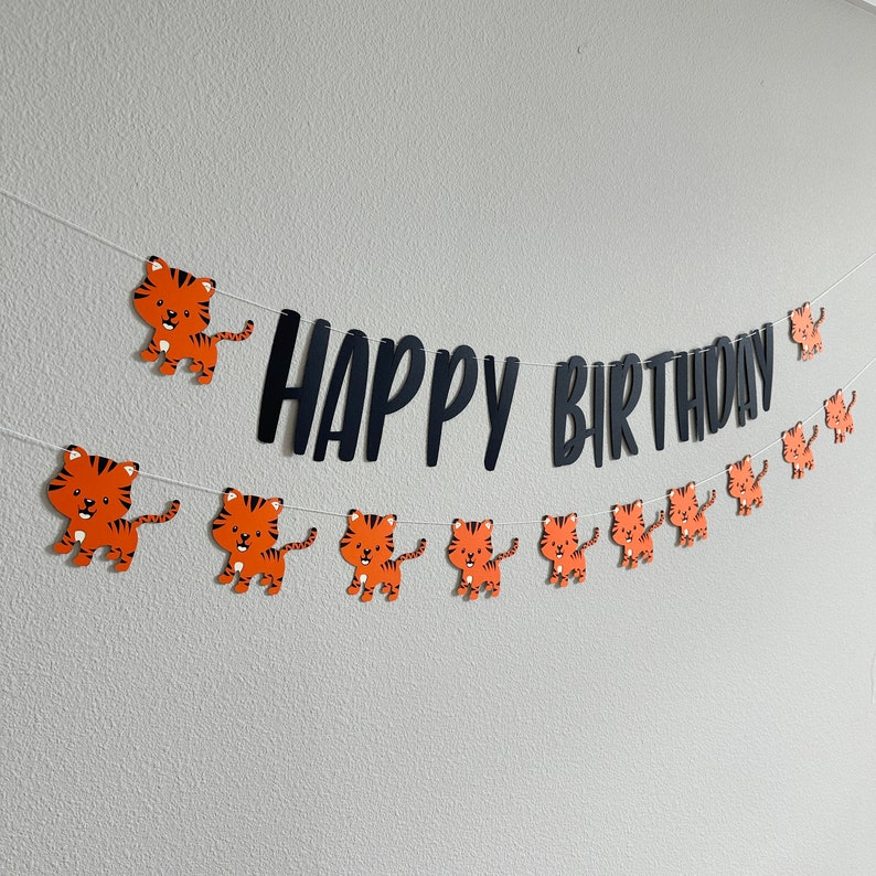 May include: A black and white banner with the text "Happy Birthday" and orange tiger cutouts. The banner is strung with white string and is hanging on a white wall.