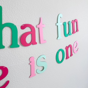 Puede incluir: Una pancarta de papel colorida con las palabras "What fun is one" en rosa, verde y azul claro. La pancarta est&aacute; enhebrada en una cuerda blanca y es perfecta para una fiesta de primer cumplea&ntilde;os.