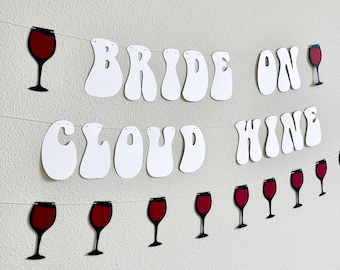 May include: A white banner with the words "BRIDE ON CLOUD WINE" in a whimsical font, strung with wine glass cutouts. The wine glasses have a red wine-filled bowl and a black stem. The banner is hanging against a white wall.
