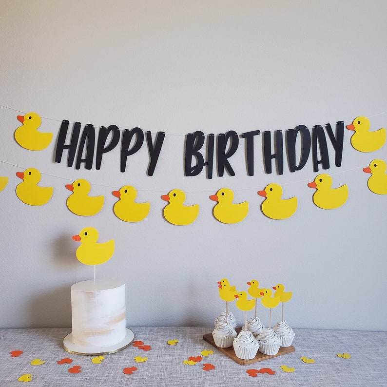 May include: A yellow rubber ducky themed birthday party with a cake decorated with a rubber ducky on top and cupcakes with rubber ducky toppers. A black banner with the text "Happy Birthday" hangs above the table.