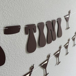 May include: A brown paper banner with the word "TINI" in large, stylized letters. The banner is strung with white string and has small brown paper martini glass cutouts hanging from it.