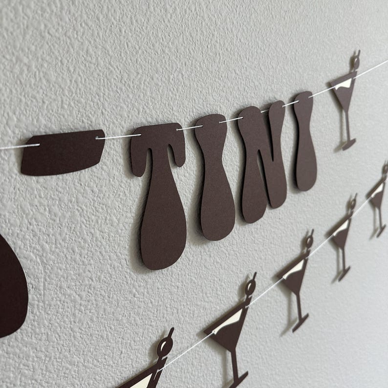 May include: A brown paper banner with the word "TINI" in large, stylized letters. The banner is strung with white string and has small brown paper martini glass cutouts hanging from it.
