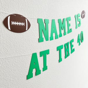 Puede incluir: Pancarta de papel verde y blanca con el texto "NAME IS AT THE 40". La pancarta est&aacute; decorada con balones de f&uacute;tbol marrones.