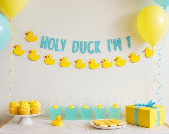 May include: A festive birthday party scene with yellow balloons, cupcakes, and a banner that reads "HOLY DUCK I'M 1." The table is set with blue drinks, rubber duckies, and a wrapped gift. A fun, celebratory theme.