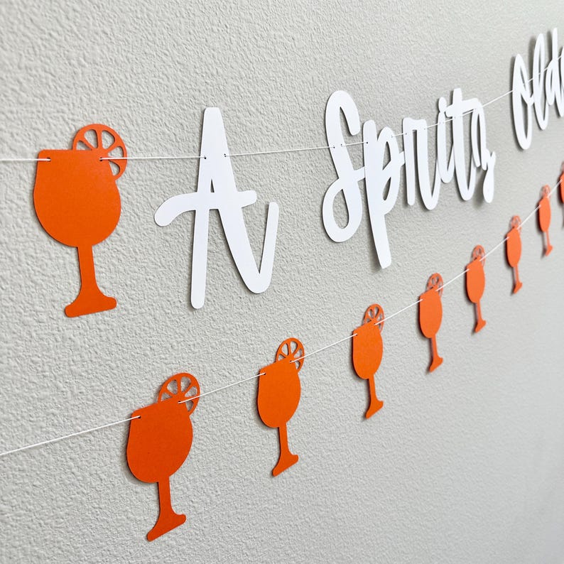 May include: A white banner with the text "A Spritz Old Fashioned" hanging from a string with orange paper cutouts of a cocktail glass with a lemon slice.