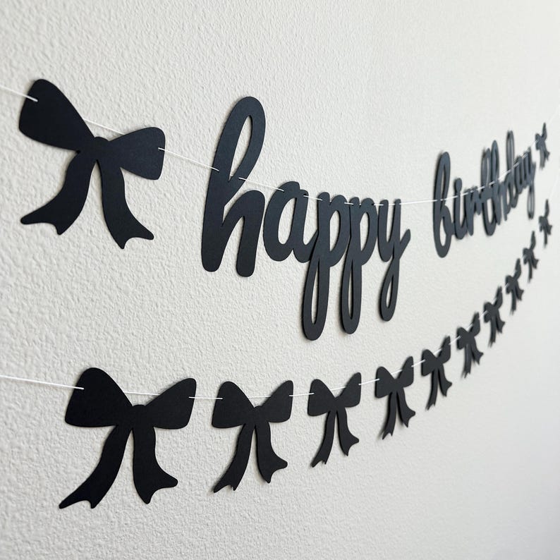 Pode incluir: Faixa de papel preta e branca com as palavras "feliz anivers&aacute;rio" e la&ccedil;os de papel preto enfiados em um barbante branco.