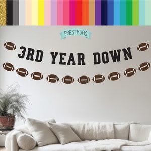 May include: A black and white banner with the text "3rd Year Down" and football-shaped cutouts. The banner is strung on a white string and is hanging on a white wall. The banner is pre-strung.