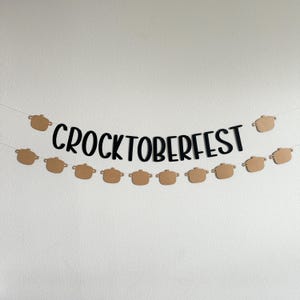 May include: A banner with the text "CROCKTOBERFEST" in black, strung between two strings. Below the text are several small, brown pot-shaped cutouts. The banner is against a white wall.