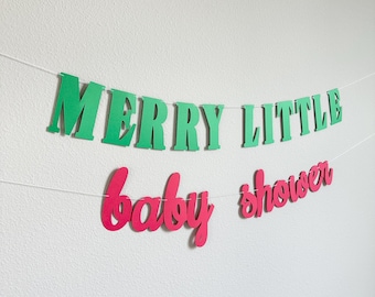 May include: A festive banner with the words "MERRY LITTLE" in green block letters and "baby shower" in pink cursive letters. The banner is strung on a white string and is against a white wall.
