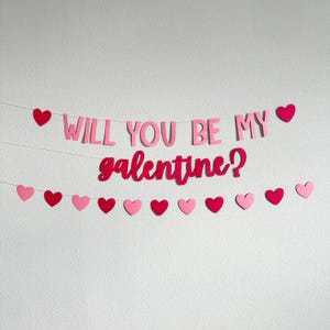 May include: A pink and red banner that says "WILL YOU BE MY galentine?" with heart-shaped decorations. The banner is strung across a white wall, with a mix of pink and red hearts.