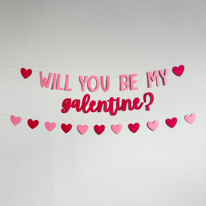 May include: A pink and red banner that says "WILL YOU BE MY galentine?" with heart-shaped decorations. The banner is strung across a white wall, with a mix of pink and red hearts.