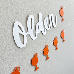 May include: White paper banner with the word "older" in cursive script. A string of orange paper cutouts of mimosa glasses with orange slices are strung below the banner.