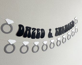 Może przedstawiać: Czarno-biały papierowy baner z tekstem "Dazed & Engaged" w retro czcionce. Baner jest ozdobiony białymi diamentowymi pierścieniami i szarymi wycięciami pierścieni.
