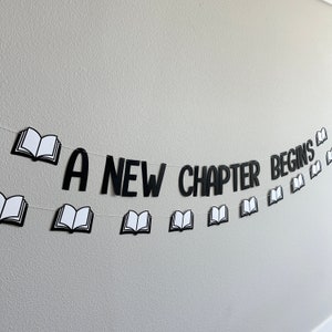 May include: A black and white banner with the text "A New Chapter Begins" and open book illustrations. The banner is strung on a white string.