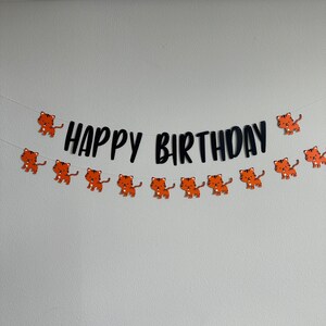 May include: A black and white happy birthday banner with orange tiger illustrations. The banner is strung with white string and is hanging on a white wall.