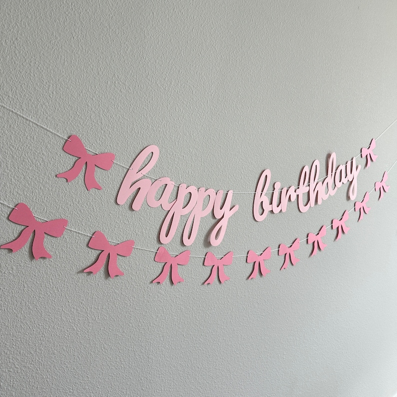 May include: Pink paper banner with the words "happy birthday" and pink bows strung on a white string.