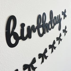 Pode incluir: Faixa de anivers&aacute;rio preta e branca com a palavra "anivers&aacute;rio" em letras grandes e cursivas e uma sequ&ecirc;ncia de la&ccedil;os pretos.