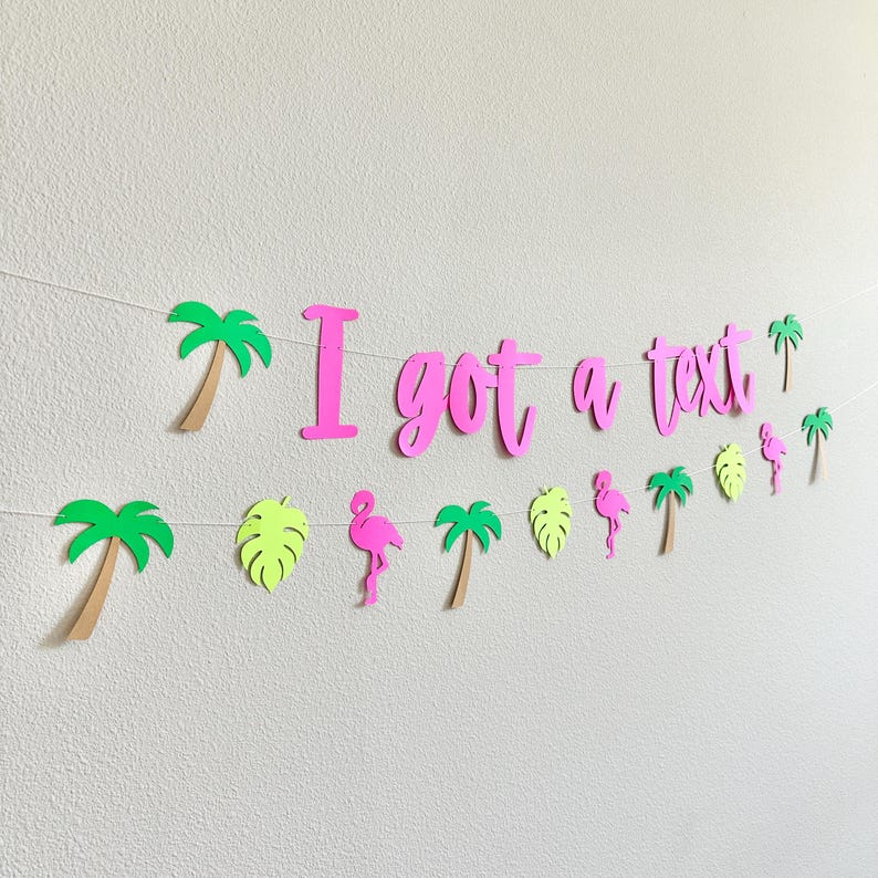 Puede incluir: Una guirnalda decorativa con la frase "I got a text" en rosa brillante, adornada con recortes de palmeras, flamencos y hojas. Las palmeras tienen hojas verdes y troncos marrones. El flamenco es rosa y las hojas son de color verde claro.