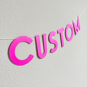 May include: A bright pink paper banner spelling the word CUSTOM in capital letters. Each individual letter is cut out and strung together on a thin white string, creating a horizontal display of the text against a plain light background.