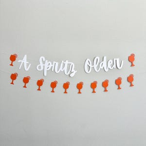 May include: A white paper banner with the text "A Spritz Older" and orange paper cutouts of cocktail glasses with a lemon slice.