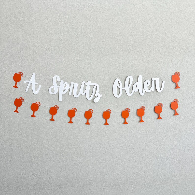 May include: A white paper banner with the text "A Spritz Older" and orange paper cutouts of cocktail glasses with a lemon slice.