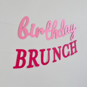 May include: Pink and red paper banner with the text "birthday brunch" hanging on a white wall.