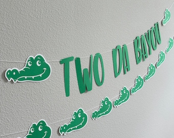 May include: Green alligator-shaped paper cutouts with the words "TWO DA BAYOU" strung together on a white string. This is a party banner for a child's second birthday.