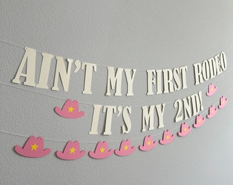 Könnte beinhalten: Weißes Banner mit dem Text "Ain't my first rodeo, it's my 2nd!" mit rosa Cowboyhüten mit gelben Sternen, die daran hängen.