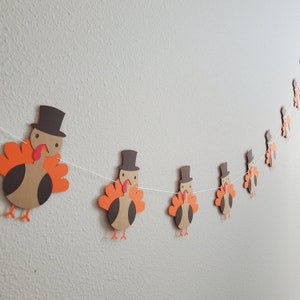 May include: A garland of brown and orange paper turkeys with black eyes and red beaks. Each turkey is wearing a brown top hat. The garland is strung on white thread.