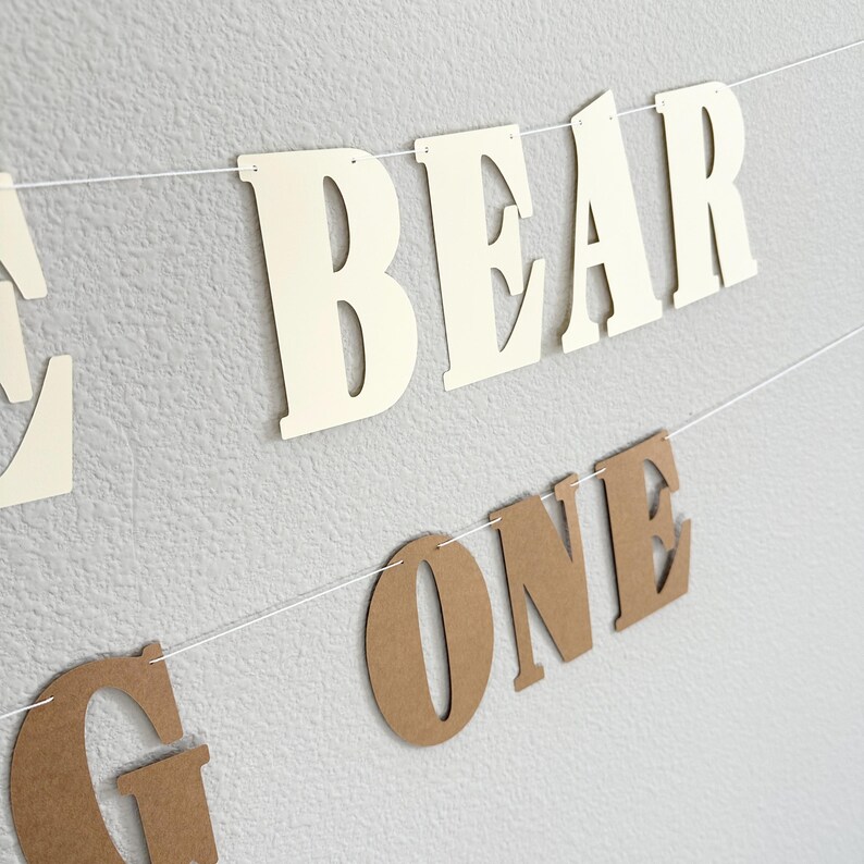 May include: A banner with the words "BEAR ONE" in cream and brown letters strung on a white string. The banner is hanging against a textured, light gray wall. The letters are cut out.