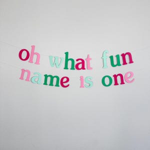 Puede incluir: Una pancarta de papel colorida con el texto "oh what fun name is one" en letras rosas, verdes y azul claro. La pancarta est&aacute; enhebrada en una cuerda blanca y est&aacute; colgada en una pared blanca.