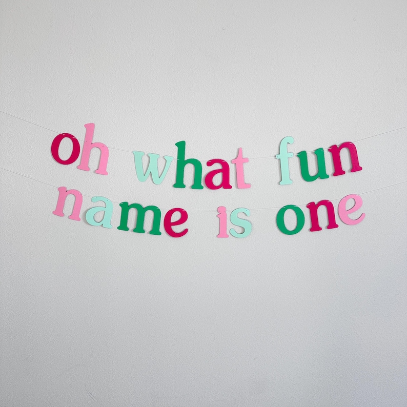 Puede incluir: Una pancarta de papel colorida con el texto "oh what fun name is one" en letras rosas, verdes y azul claro. La pancarta est&aacute; enhebrada en una cuerda blanca y est&aacute; colgada en una pared blanca.