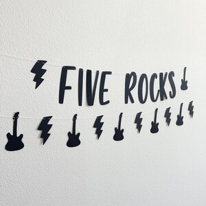 May include: Black banner with the words "FIVE ROCKS" and guitar and lightning bolt cutouts. The banner is strung on a white string and is against a white wall. The banner is a rock and roll themed decoration.